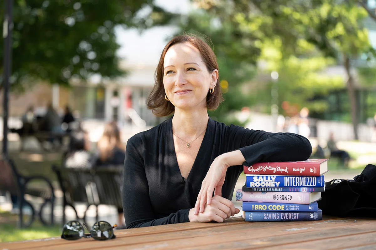 Author and Carleton University professor Sarah Brouillette sits in Carleton's quad with several books stacked beside them. Visible titles include "Conversations with Friends" by Sally Rooney, "Intermezzo," "Big Boned," "The Bro Code," and "Saving Everest." The background shows a park-like setting with benches and trees.