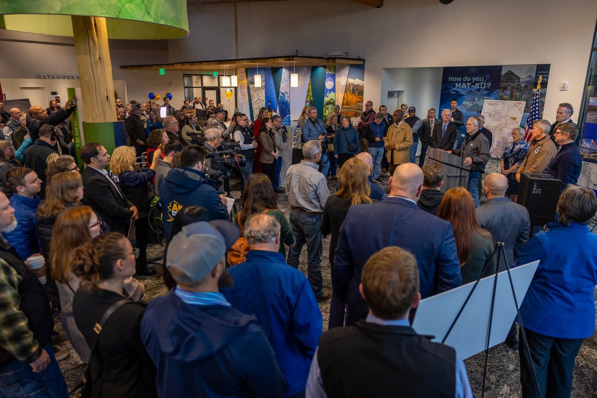 A large crowd gathers indoors for a formal presentation or public event. Several people stand near a podium while attendees watch; cameras and display boards are visible. A wall poster contains text.