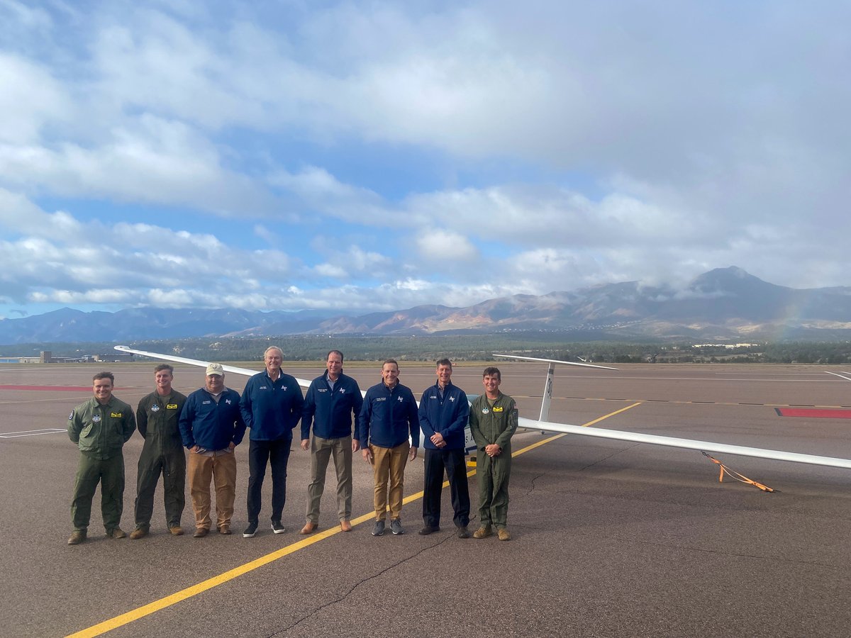 A group of eight men pose on an airport runway in front of a glider, with mountains and a cloudy sky in the background. Some clothing and the aircraft have visible text or logos.