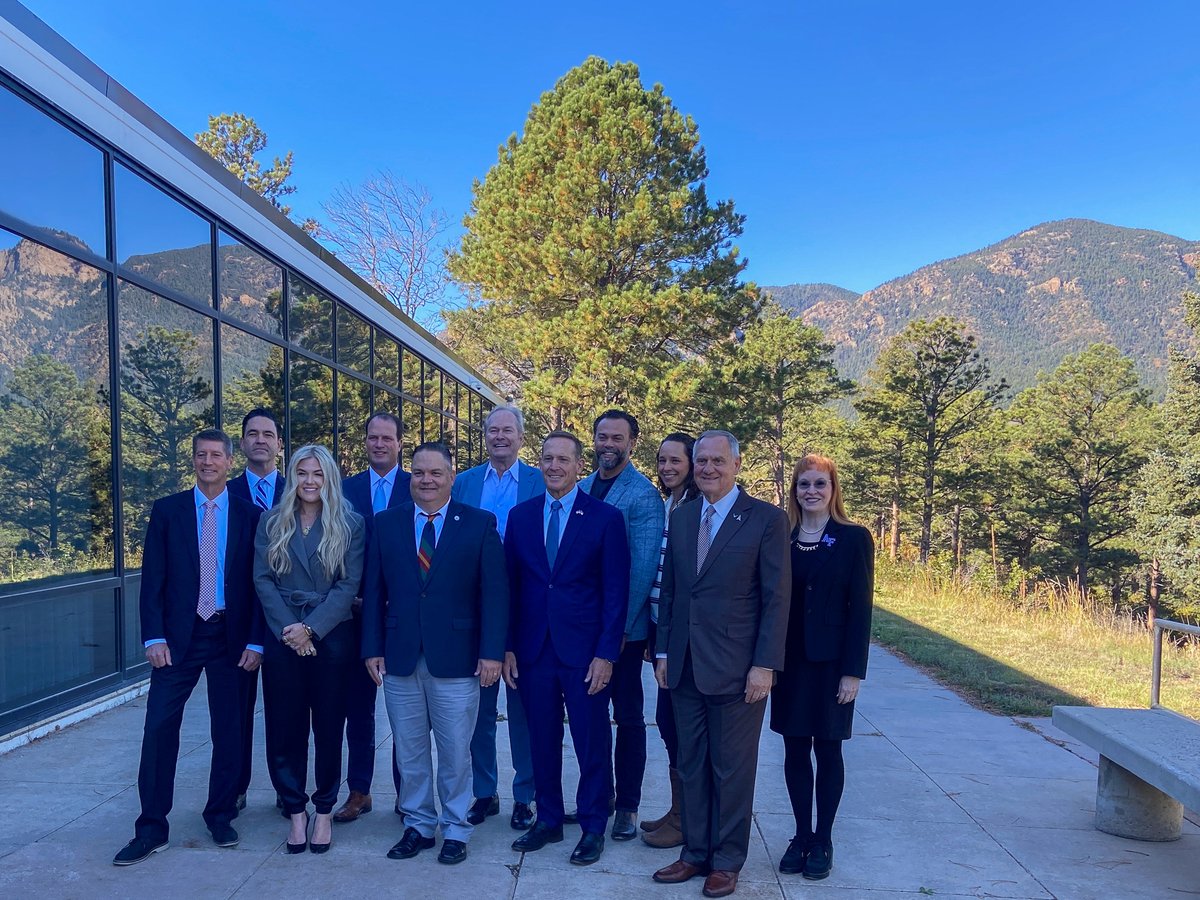 A group of ten adults pose together outdoors beside a modern glass building, with pine trees and mountainous scenery in the background. Some clothing has small visible text or logos.
