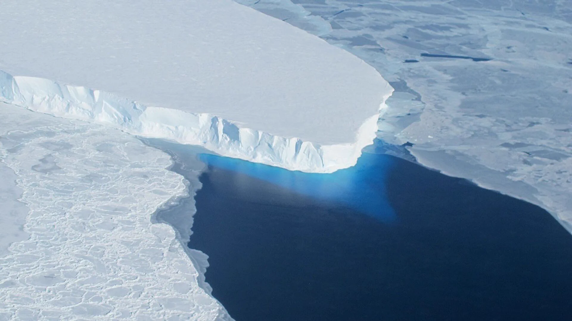 Aerial view of Thwaites Glacier in Antarctica, showing a vast expanse of white ice ending in a steep ice cliff above dark blue ocean water. Broken ice surrounds the glacier’s edge. Credit: NASA