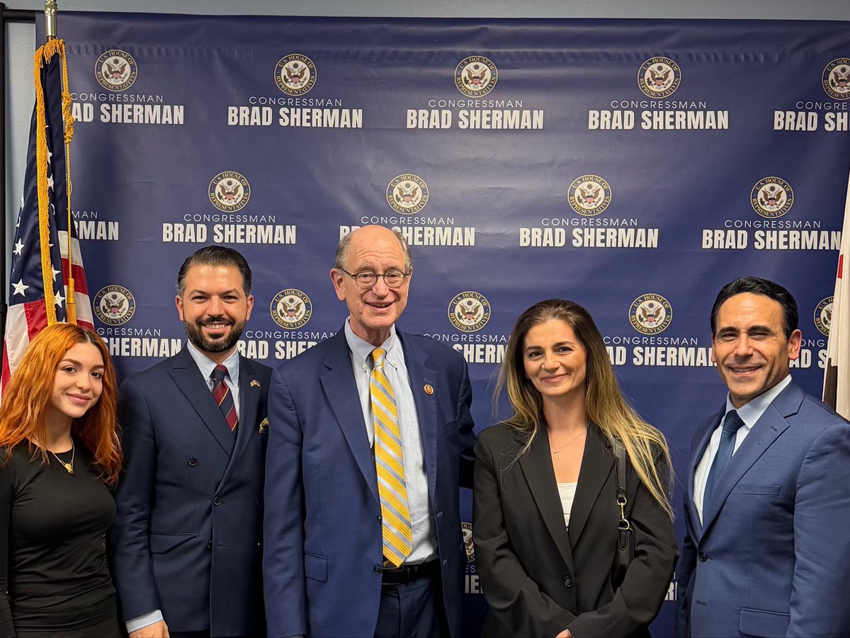 Five people pose for a group photo in front of a blue congressional backdrop. An American flag is visible at the left, and text and emblems repeat across the backdrop.