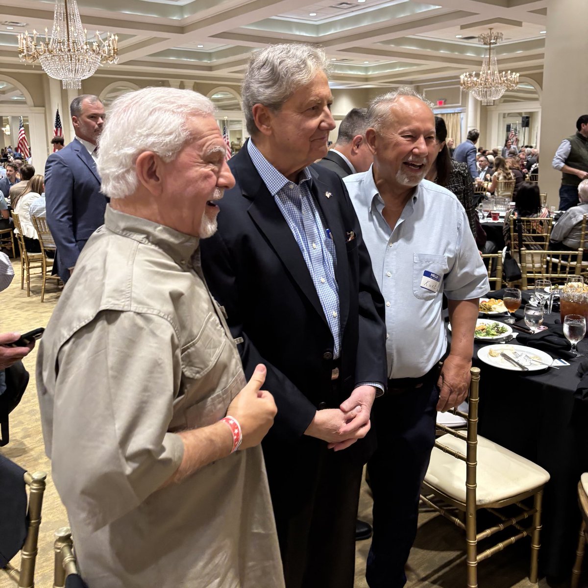 Three men pose together and smile at a formal banquet in a large ballroom. Guests, tables, chairs, chandeliers, and flags are visible in the background. One man wears a name badge with text.