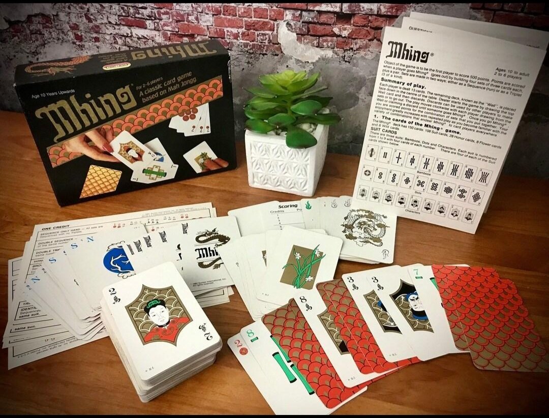 Mastermonkdaddy's tweet photo. Have you ever seen this card game before?

This was my family's favorite game to play on game night!

Mhing was released in 1982 and the best way to describe it is a card game that's kind of a cross between Mahjong and Phase 10 using a point system. You are rewarded points for different sets that you place down out of your hand and it's a race to 500 points. 

Like Phase 10 everyone is dealt a certain amount of cards (13), with the remaining cards left in the middle along with a discard pile. You make 'sets' in your hand by matching same suits and making runs of numbered cards.

 Play begins by taking 1 card either from the discard pile or from one of the face down pile. You then discard 1 card from your hand the play goes to the next person on your left. On your turn you can lay down your sets then are allowed to play your remaining cards onto others sets that other players had laid down. You get points depending on how many in your set you were able to lay down, and you "go out" by getting rid of all the remaining cards in your hand after you completed your sets. 

This game is players 10 and up as it can be a bit difficult and each game takes about an hour to finish.

Note  *I typed this up from memory so if anything I said is wrong feel free to correct me