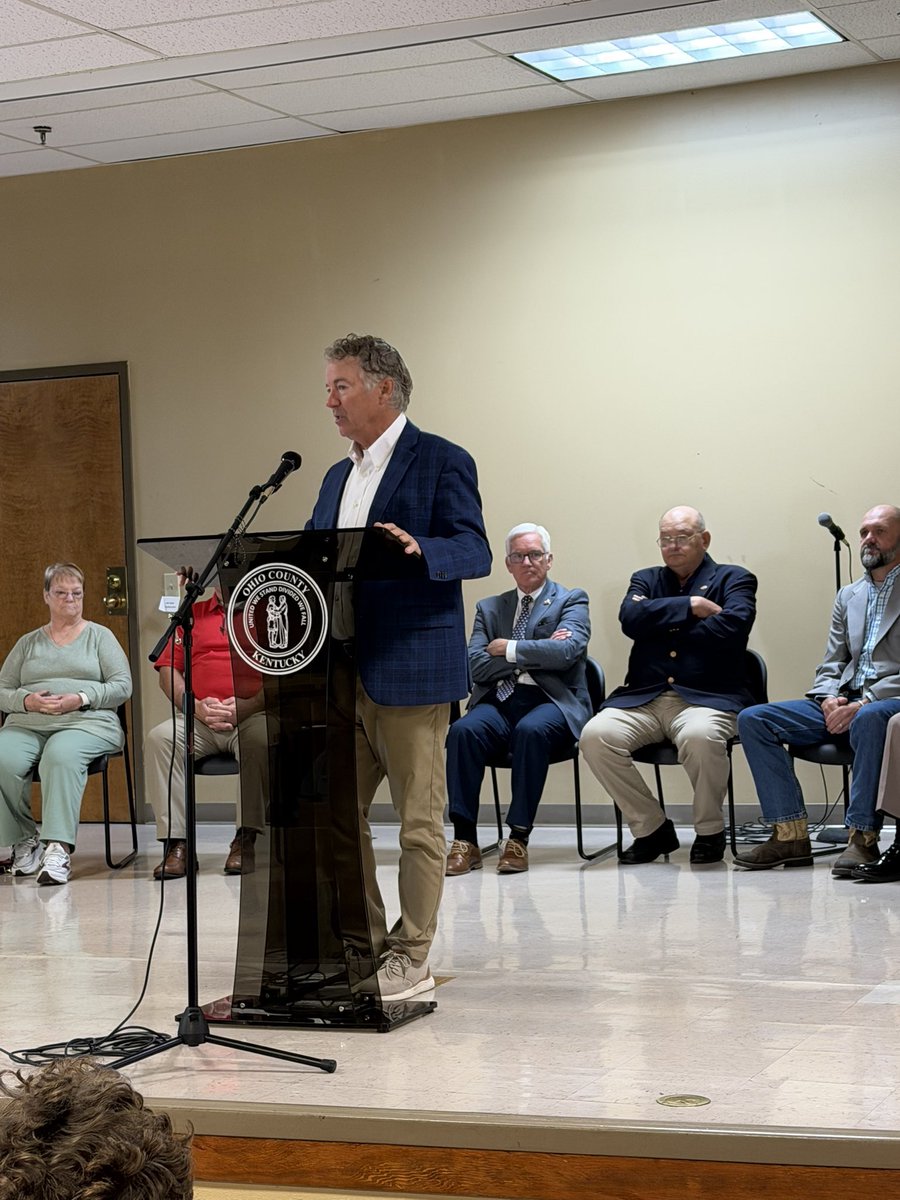A man speaks at a clear lectern during an indoor gathering, with several people seated behind him. The lectern bears a county seal, and there is text on it.