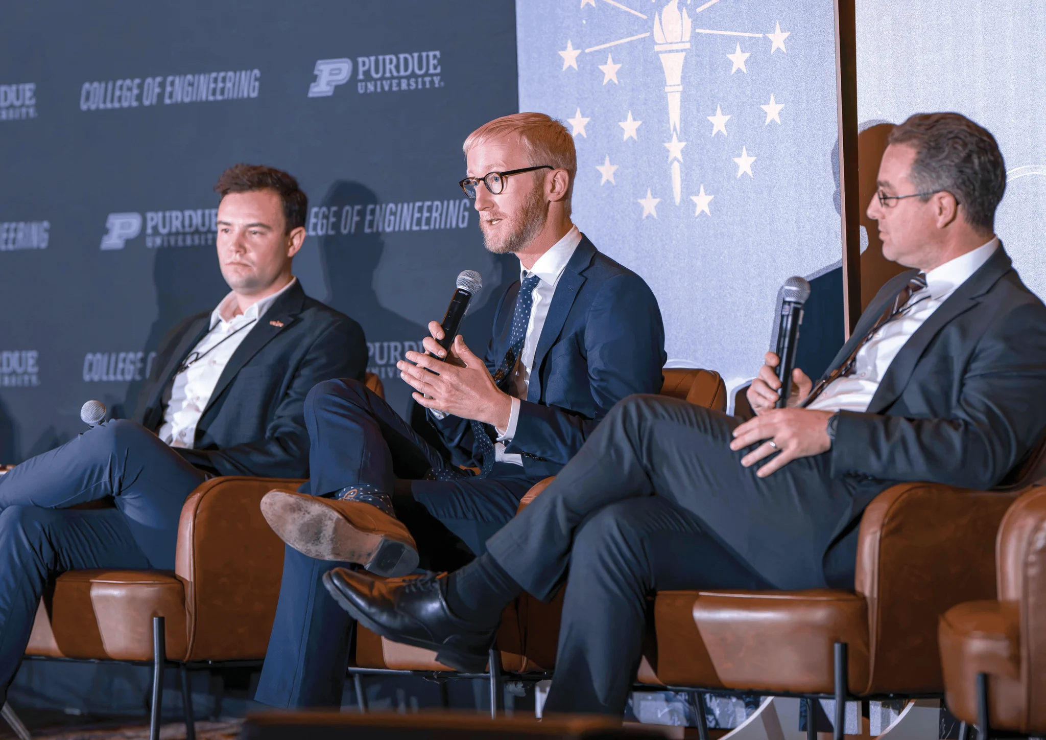 Three individuals are seated on a stage during an event at Purdue University College of Engineering. One person is speaking into a microphone. They are all dressed in formal attire. The backdrop displays the Purdue University logo and a design resembling a lighthouse.
