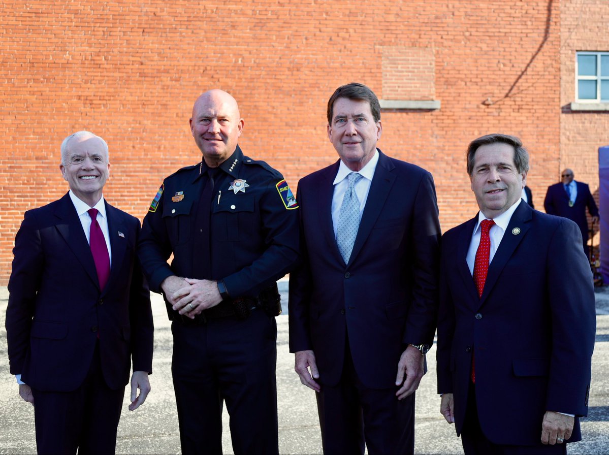Four men pose together outdoors in front of a brick building. One wears a police uniform, while the others wear business suits. A fifth person is visible in the background. There is a small amount of text on the uniform and clothing.
