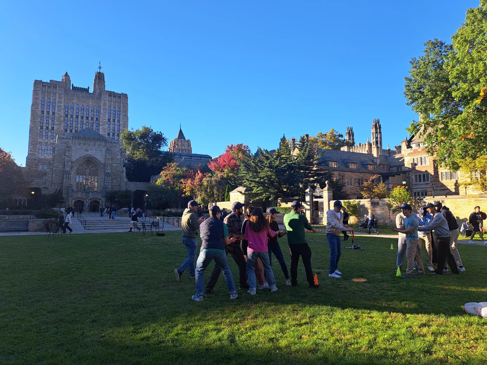A group of people are participating in an outdoor activity on a grassy area at Yale University. The background features historic campus buildings under a clear blue sky.