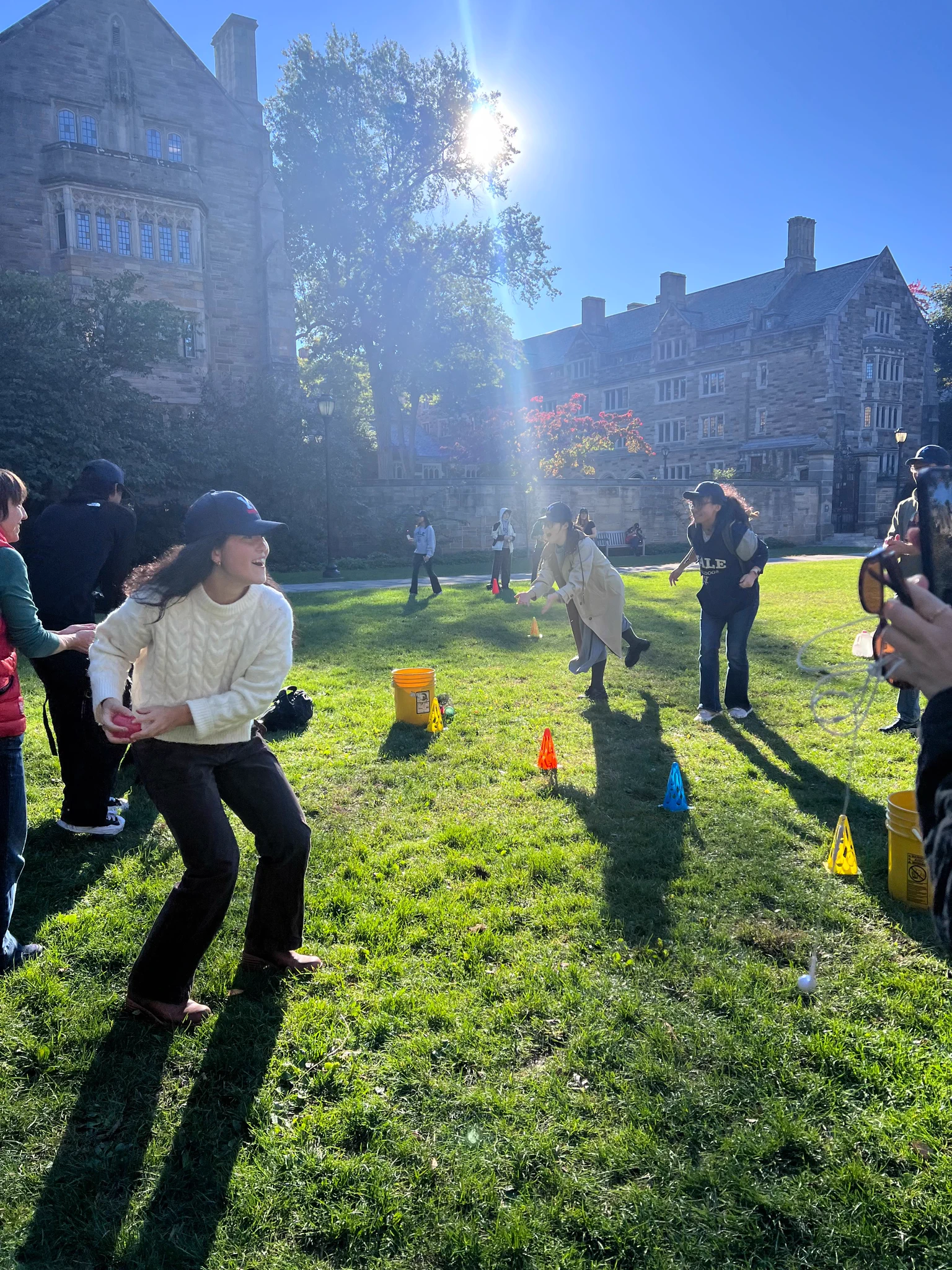 People are gathered outdoors on a grassy area with trees and a stone building in the background. It's a sunny day, and there are colorful cones and equipment on the grass.