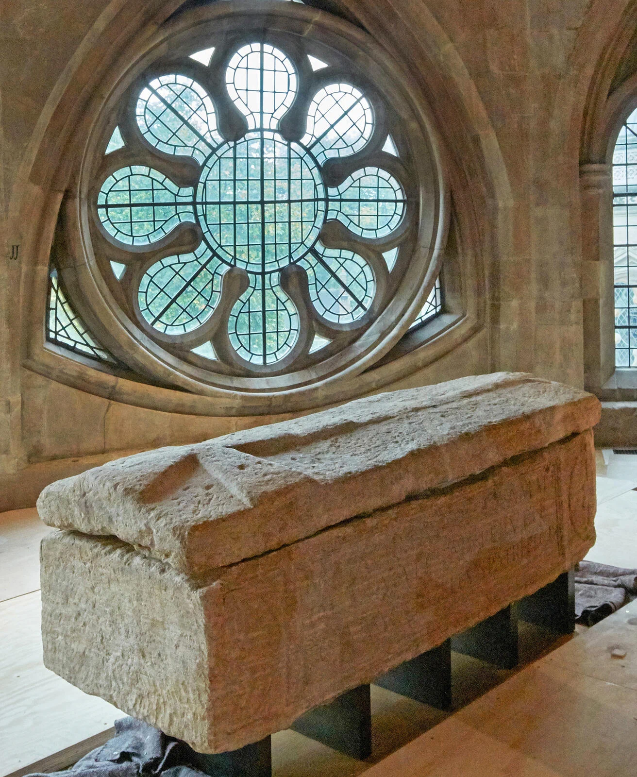 A stone sarcophagus rests indoors on metal supports. Behind it, a large, ornate circular rose window with geometric stone tracery is set into a tan stone wall.\n(Image credit to Dean & Chapter of Westminster)