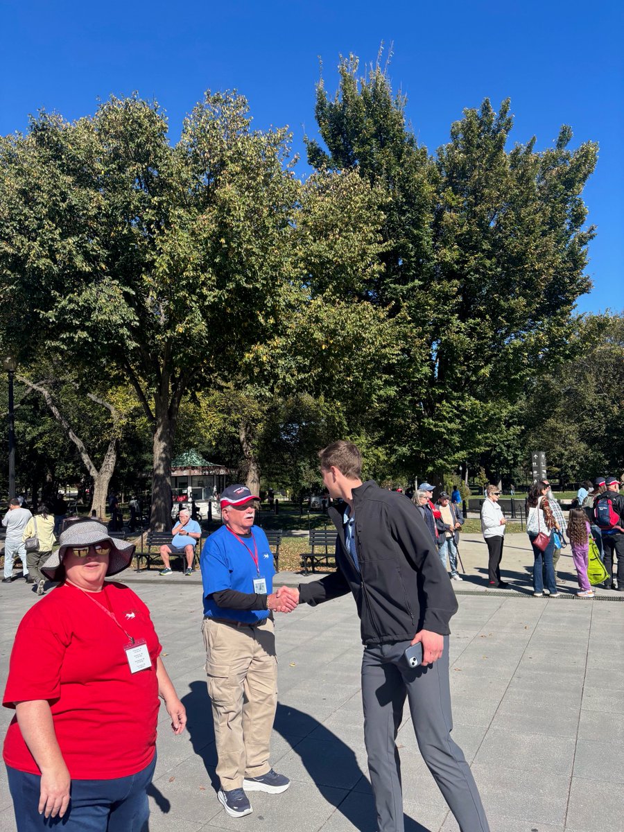 A sunny outdoor park plaza with several people gathered. In the foreground, two men are shaking hands while a woman stands nearby; other visitors, trees, benches, and a small kiosk are visible in the background. Some clothing and signs contain text.