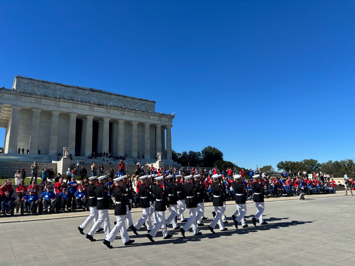 A group of uniformed military personnel marches in formation across a plaza in front of a large neoclassical memorial, while spectators watch from the steps and along the sides. There is some text on the building.