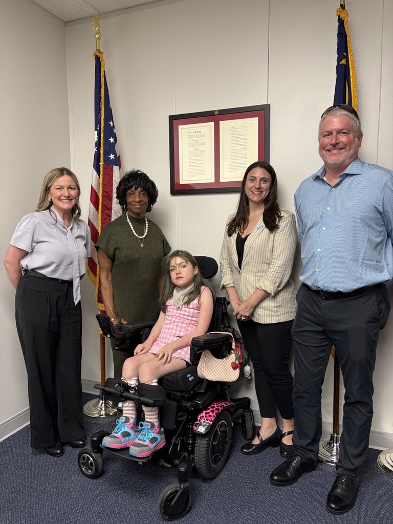 A group of five people pose together in an office, with a young girl seated in a motorized wheelchair. Two flags and a framed document are visible behind them. There is text on the document.