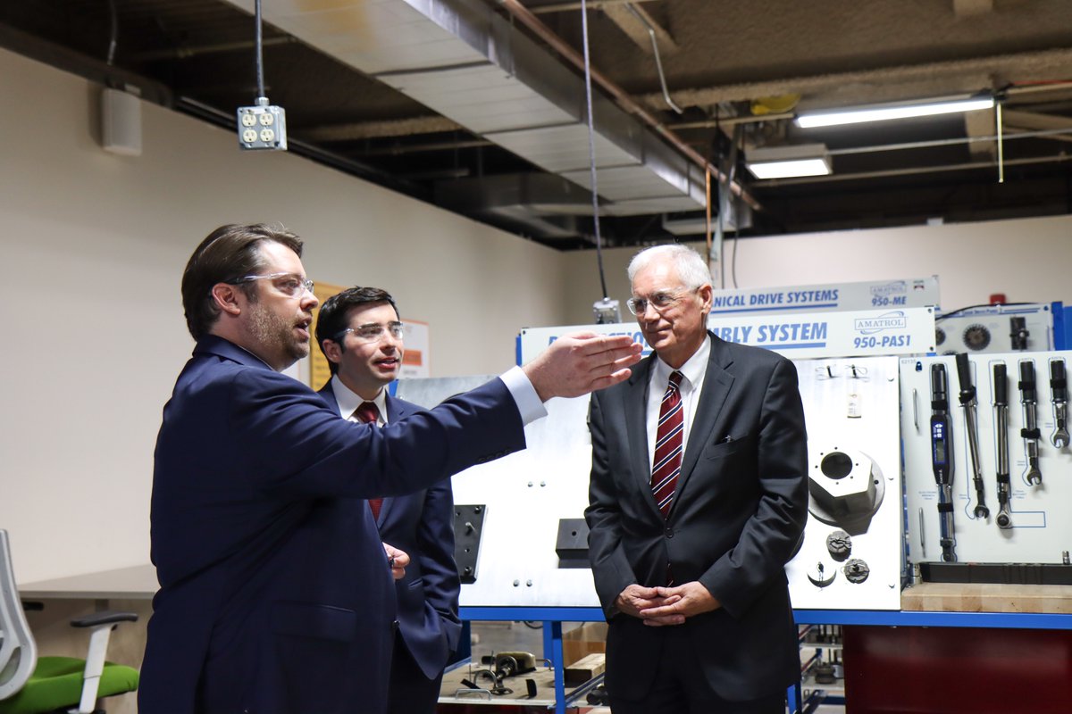 Three men in suits stand in a technical training workshop. One man gestures toward equipment while the others listen. There is text on signs attached to the machinery.