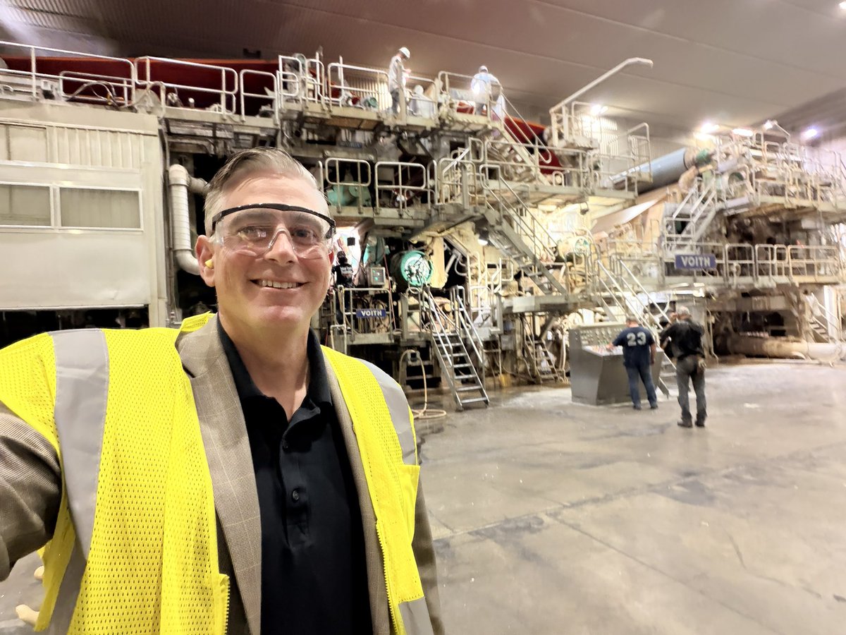 A man wearing safety glasses and a bright yellow safety vest takes a selfie inside a large industrial facility. Complex machinery, elevated walkways, railings, stairs, and several workers are visible in the background. There is text on some equipment and clothing.