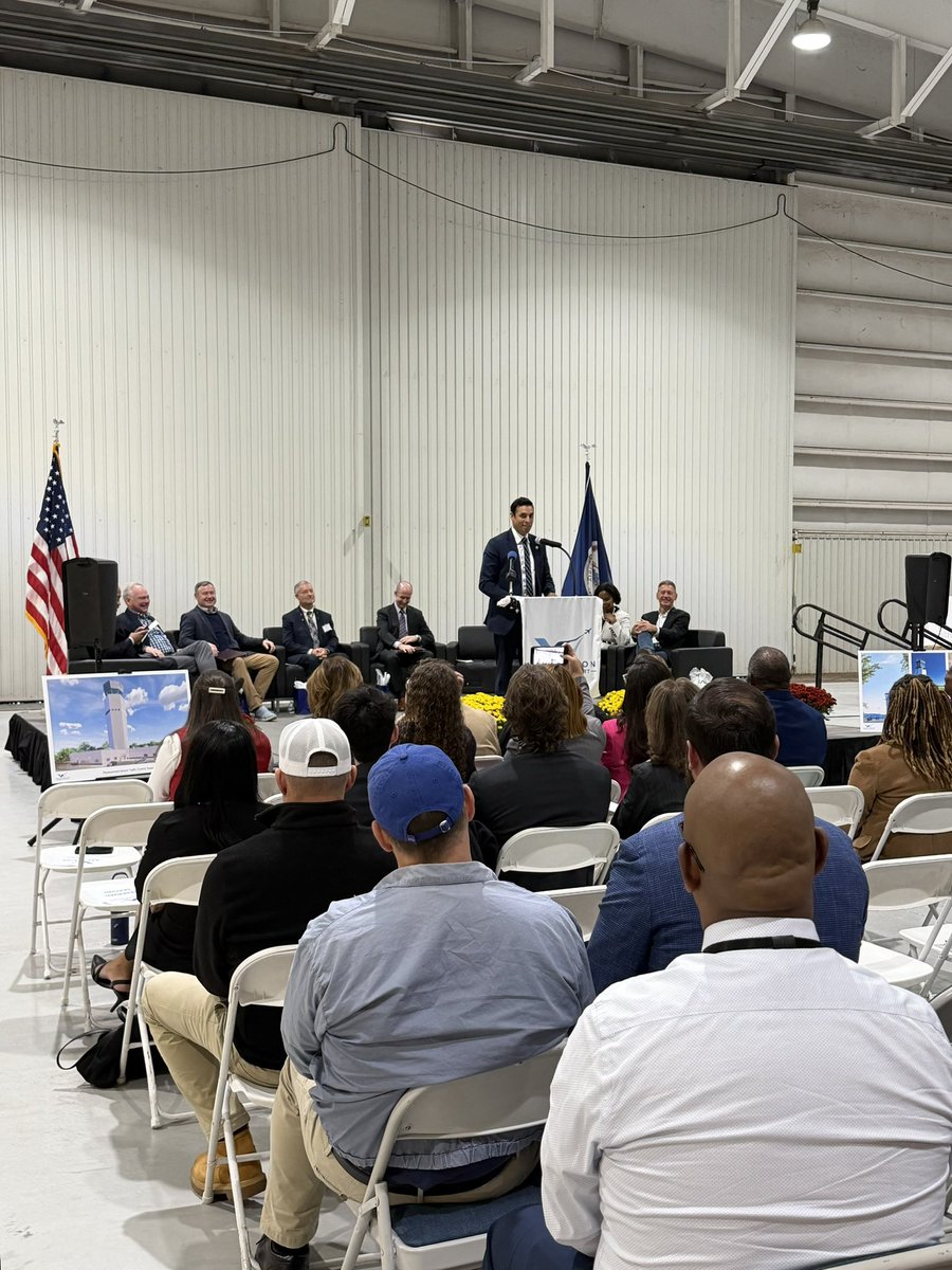 A man speaks at a podium during an indoor public event while several people sit on stage and an audience watches from rows of folding chairs. Flags, speakers, flower arrangements, and display boards are visible. Some text appears on the podium and boards.