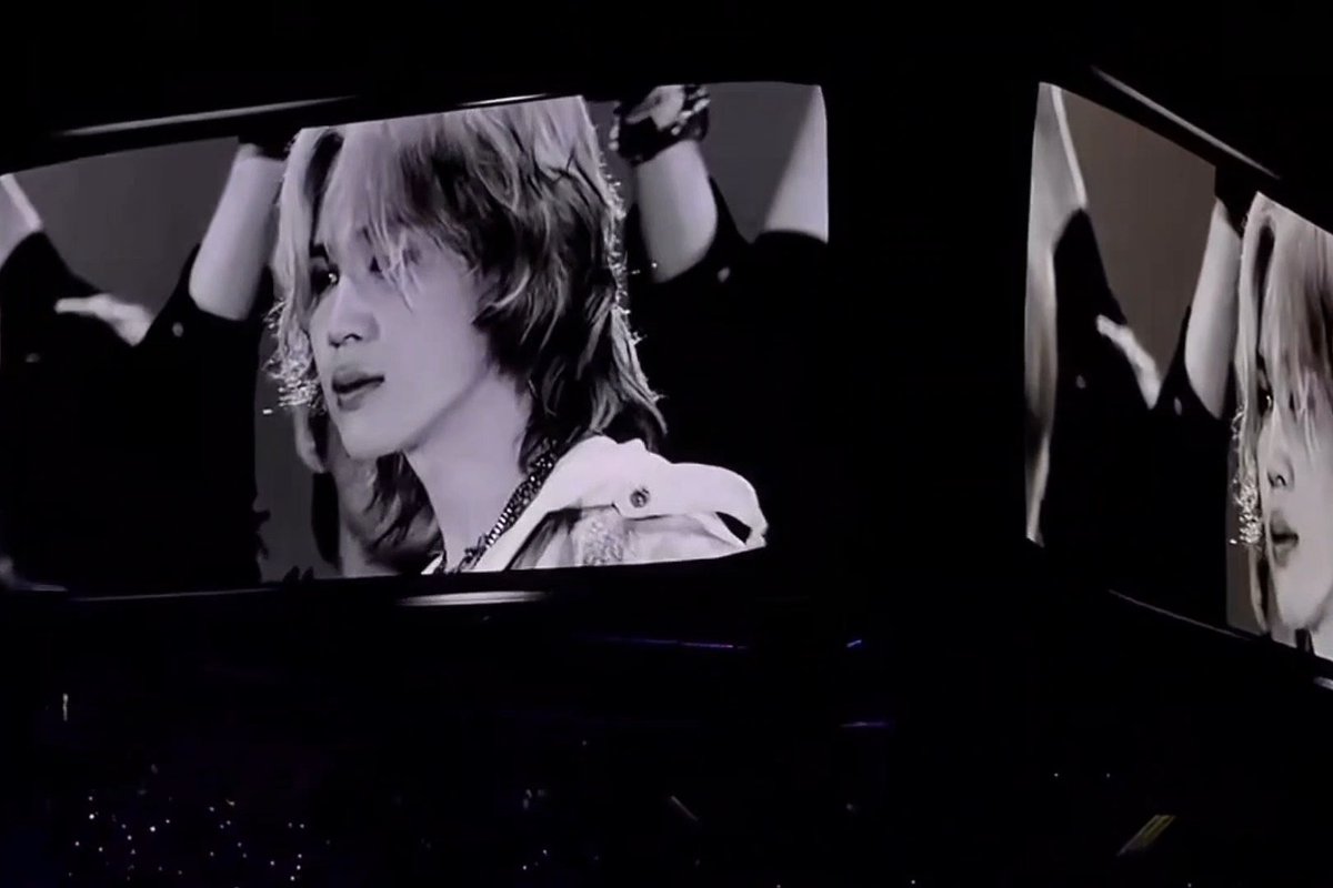 A black-and-white concert close-up shows a shaggy-haired male performer on a large curved stage screen, with his face repeated on a second screen to the right.
