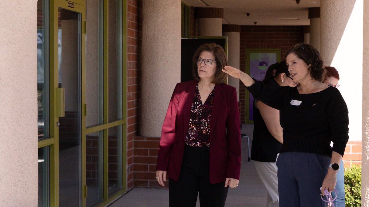 Several women stand in a covered outdoor corridor beside a building. One woman gestures toward a glass doorway while another looks in that direction. There is text on the doorway and on a name badge.