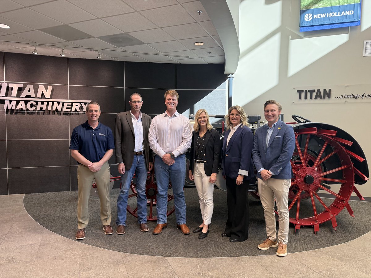A group of six adults pose for a photo inside a machinery showroom, standing in front of large red agricultural equipment. Company signs are visible on the walls.