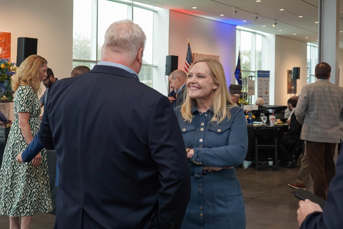 A group of people mingle and talk at an indoor reception or event. A smiling blonde woman in a blue outfit speaks with a man in a dark suit, while other attendees stand or sit around tables. Flags, speakers, large windows, and event displays are visible in the background. There is some text on a display.