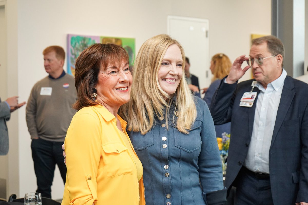 A photograph of two smiling women posing together at an indoor gathering, with several people in the background. Some people are wearing name badges, and there is artwork on the wall. Text appears on badges and signs.