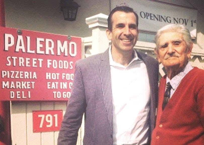 Two men pose together outside a storefront. A red sign advertises a food business, and another sign above mentions an opening date. Text is visible throughout the scene.