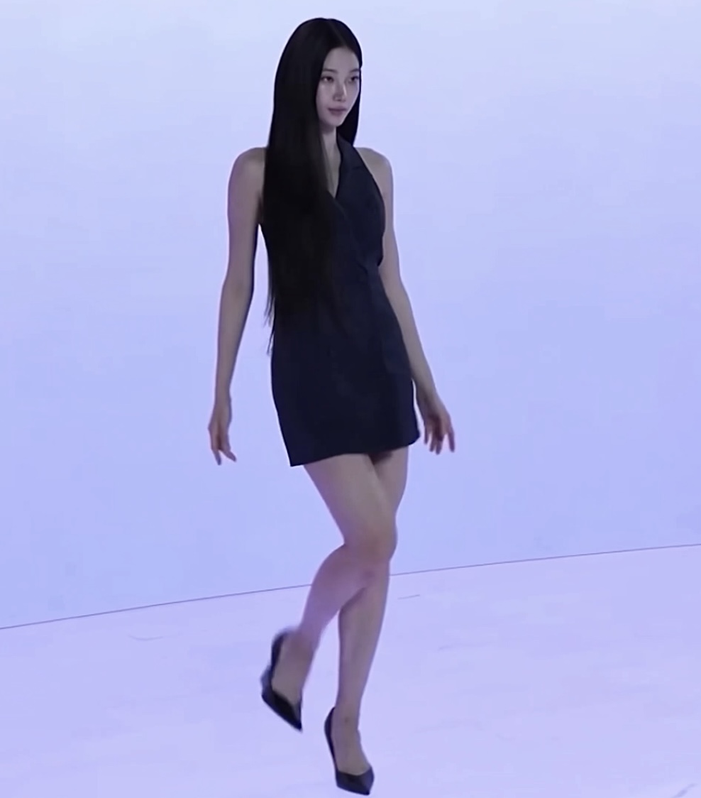 A woman with very long black hair walks across a pale, minimalist studio or runway space wearing a short black halter dress and black pointed high heels.
