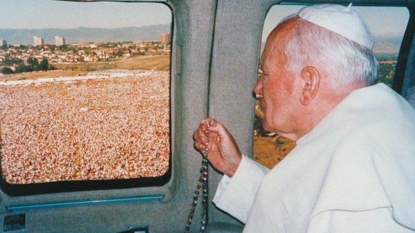 A photograph shows an elderly man in white religious clothing looking out of a vehicle window at a large crowd gathered in an open field. He holds a string of beads. There is text on the image.