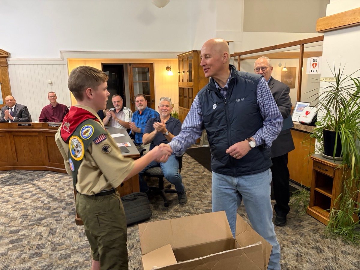 A Boy Scout shakes hands with a man in a municipal meeting room while several seated adults applaud. An open cardboard box is in the foreground. There are a few signs and nameplates with text.