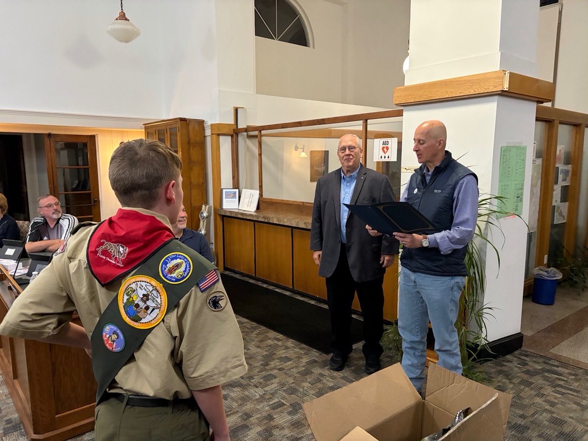 A scout in uniform stands facing two men in a community room. One man holds an open folder while the other stands beside him. Other people sit at a table in the background, and an open cardboard box is on the floor. There is some visible text on signs and uniform patches.