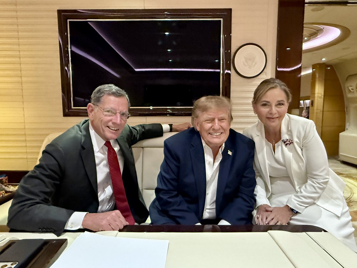 Three adults in formal clothing sit close together and smile at the camera in an upscale interior. A large dark television, a round wall decoration, and a table with papers are visible. There is no prominent readable text.