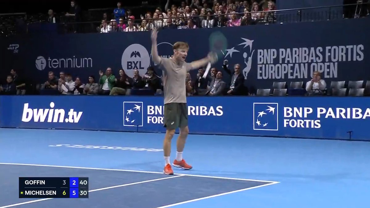 A tennis player stands on a blue indoor court at the BNP Paribas Fortis European Open, celebrating with both arms raised. The scoreboard reads “GOFFIN 3 2 40” and “MICHELSEN 6 5 30,” matching the post’s reference to David Goffin at the @BNPPFEUOpen.