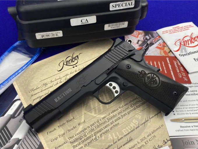 A Kimber handgun rests on a pile of papers, some branded with Kimber's logo. A black case labeled "Special" and "CA" is in the background. The image includes a "Bastion" engraving on the handgun and various promotional materials scattered around.