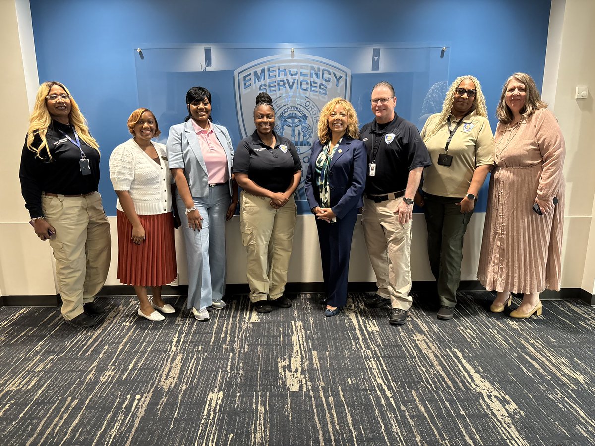 A group of eight adults pose for a photo in front of a blue wall display. Several wear uniforms or identification badges. There is text on a sign behind them.