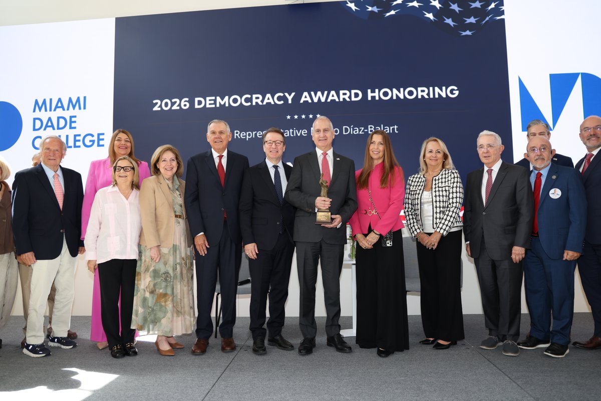 A group of adults pose for a formal event photo on a stage. One man in the center holds an award, with a large event backdrop and college branding behind them. The backdrop contains text.