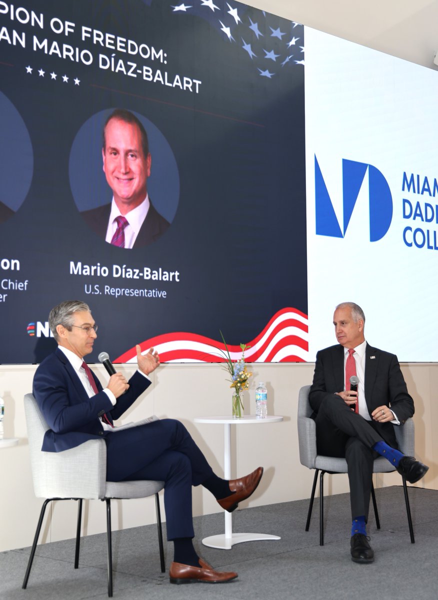 Two men in suits sit on chairs on a small stage, holding microphones during a discussion. A large display behind them shows a portrait, patriotic graphics, and text; a college logo is visible on a panel to the right.