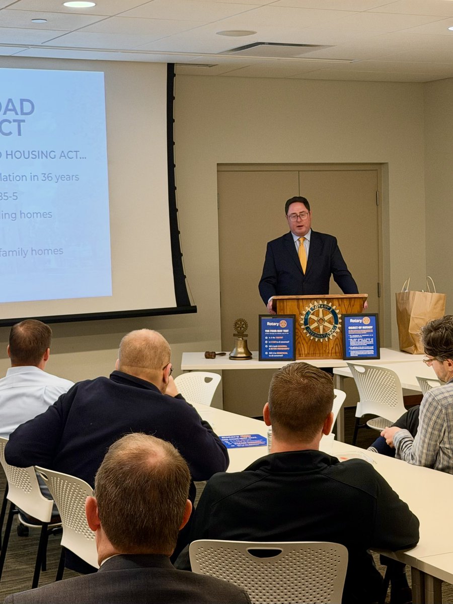 A man in a suit and yellow tie speaks from a lectern to an audience seated around tables in a meeting room. A presentation is projected on a screen, and Rotary signs and a bell sit on the lectern table. Text is visible on the projection and signs.
