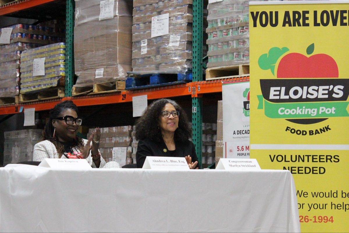 Two women sit behind a white-covered table at an event in a food-bank warehouse. Shelves of palletized goods fill the background, and a large yellow food-bank volunteer sign stands at the right. Text appears on the sign and table placards.