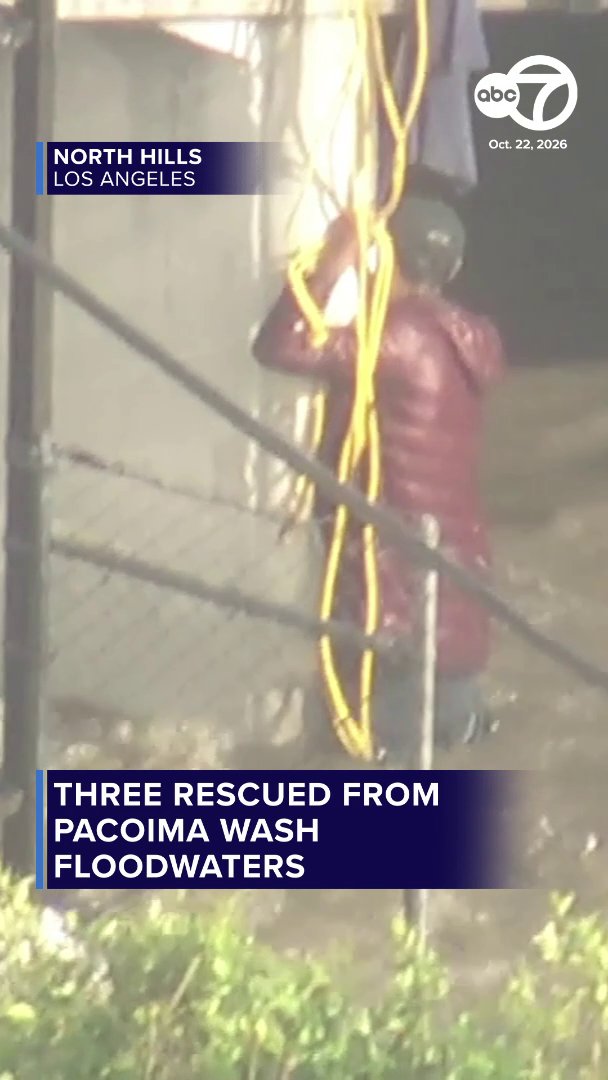 An ABC7 news image shows a rescuer in a helmet standing beside ropes along a fence in floodwater. On-screen text reads “NORTH HILLS LOS ANGELES” and “THREE RESCUED FROM PACOIMA WASH FLOODWATERS,” dated Oct. 22, 2026.