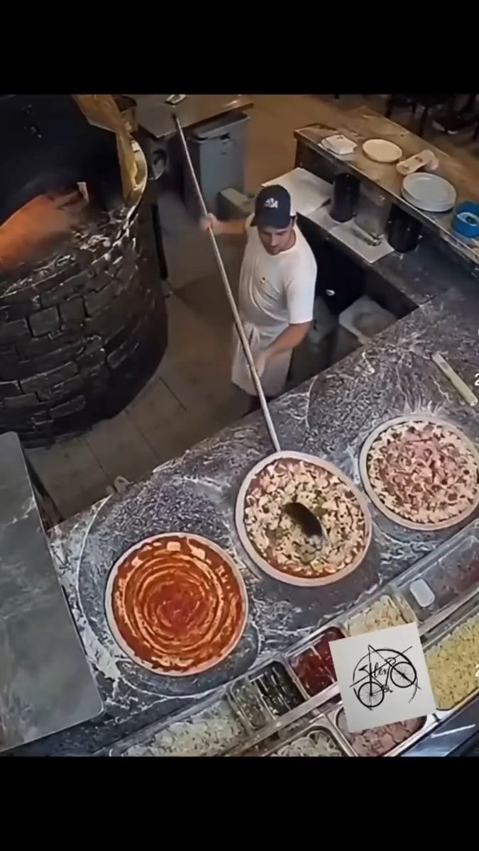 An overhead restaurant-kitchen view shows a worker using a long tool near several round food pans, including red sauce and pizza-like toppings, with a white paper marked by a black scribbled symbol.