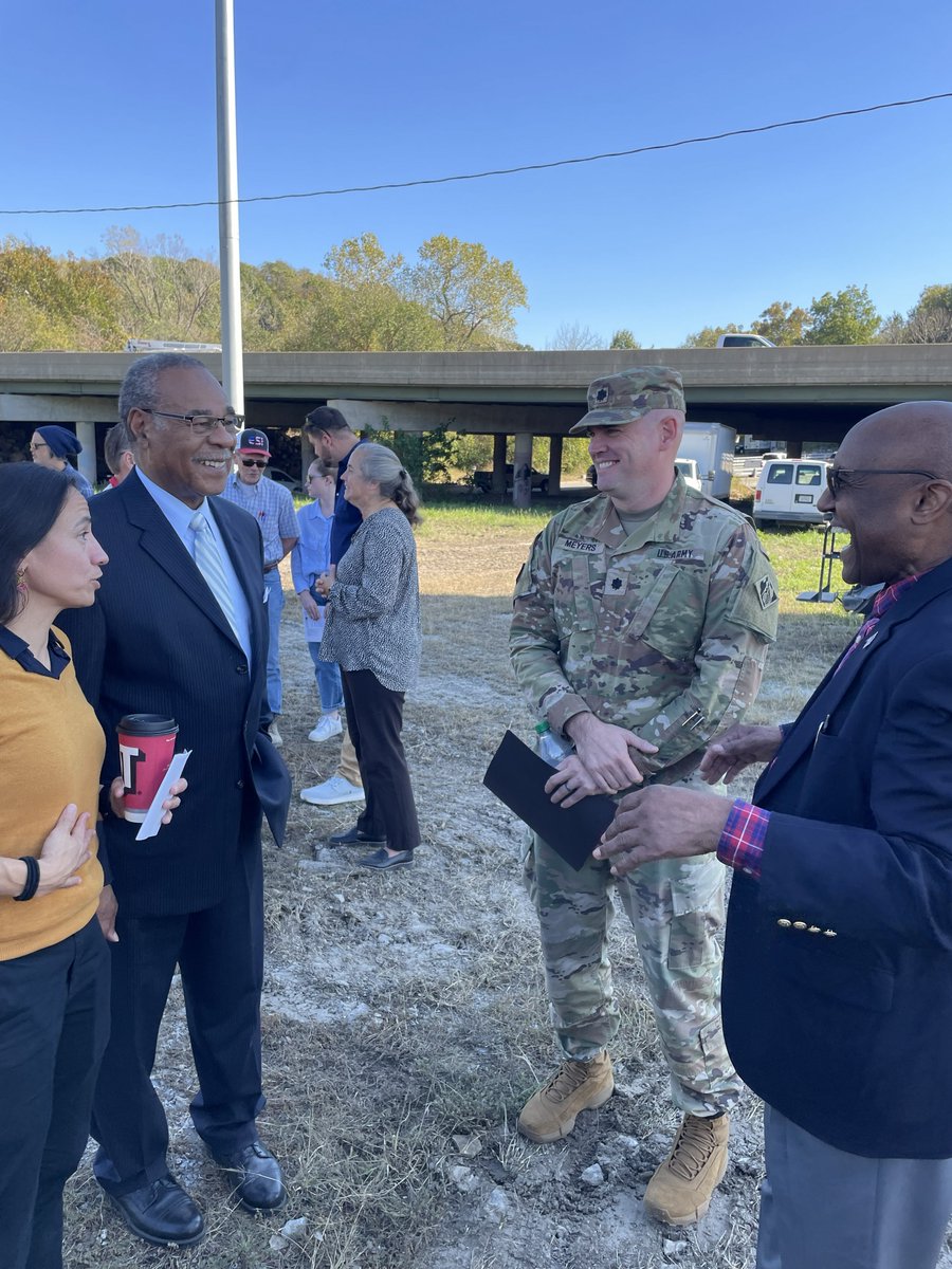 A group of people chat outdoors beside an overpass on a clear day. A soldier in camouflage stands among several civilians. Text is visible on clothing and a cup.