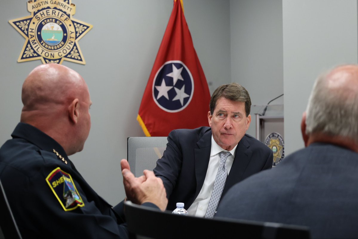 Three men sit around a table in a sheriff’s office or conference room. A uniformed sheriff speaks with a suited man while another man is seen from behind. A red state flag and sheriff’s insignia are displayed in the background, and there is visible text on the insignia.
