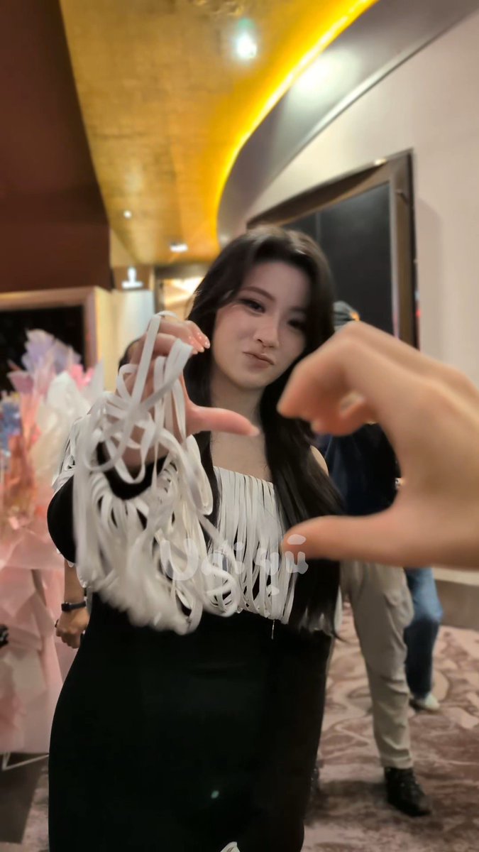 A woman in a black dress with white fringe stands indoors while making a heart shape with her hands, with hanging balloons and people visible in the background.
