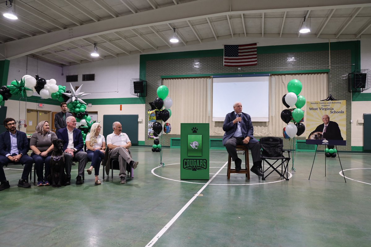 A man speaks into a microphone while seated on a stool in a school gymnasium. Several people sit nearby, and the room is decorated with balloons and an American flag. A podium and a campaign-style sign are visible. There is text on the podium and sign.
