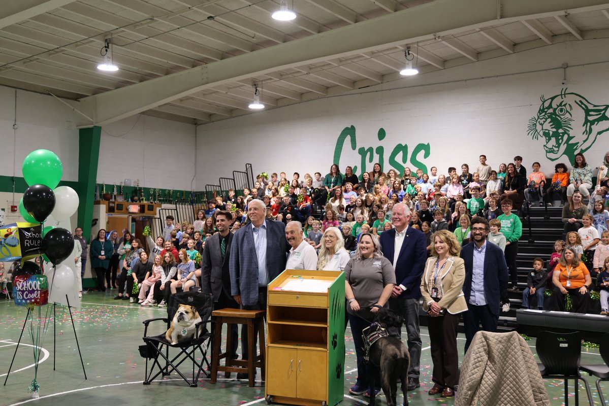 A large group of adults and children pose in a school gymnasium, with several people standing in front of packed bleachers. Two dogs are near the group, and colorful confetti, balloons, a podium, and school-themed decorations are visible. There is text on the wall and decorations.