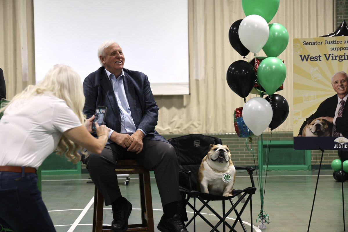 A man sits on a stool beside a bulldog seated in a folding chair while a woman photographs them. Green, black, and white balloons and a campaign-style display are visible in the background, with text on the display.