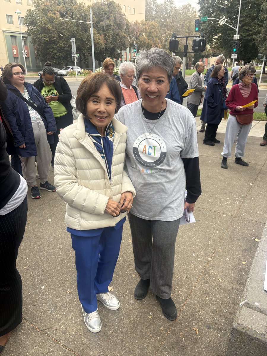 A group of people stand on a city sidewalk near an intersection. Two women pose smiling in the foreground; one wears a white puffer jacket and blue pants, and the other wears a gray graphic T-shirt. Several people behind them hold papers, with trees, buildings, traffic lights, and a street in the background. Text appears on clothing and street signs.