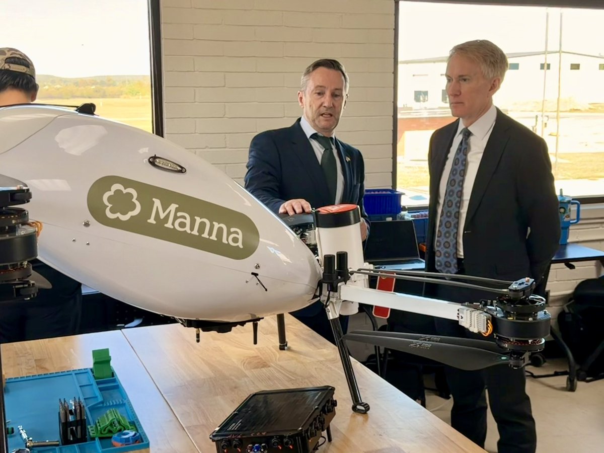 Two men in suits stand beside and inspect a large white delivery drone in an indoor workshop or demonstration space. The drone has multiple rotor arms and a branded body; tools and equipment sit on a table in the foreground. Text appears on the drone.