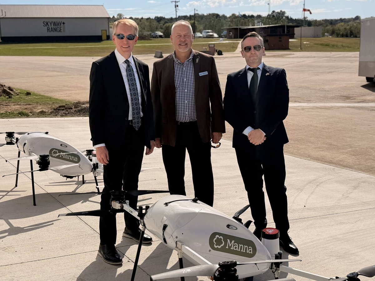 Three men in formal clothing pose on an airfield beside two white delivery drones. A hangar, buildings, parked vehicles, and open sky are visible in the background. There is text on the hangar and drones.