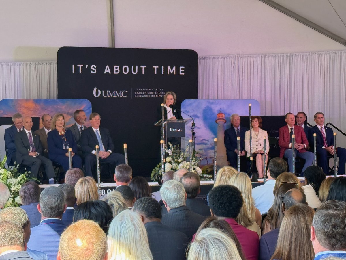 A woman speaks at a podium on a decorated stage while formally dressed guests sit behind her and an audience watches. A large backdrop displays campaign branding and text.